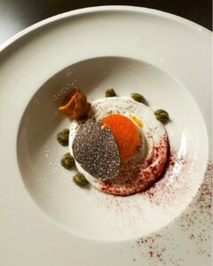 Fine-dining plate with white foam, orange yolk, black truffle slice, green purée dots, and red dust