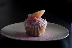 Cupcake with a swirl of pale pink frosting, a gummy fruit wedge, and pink sugar crystals on a white plate against a dark background