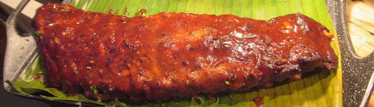 Rack of glossy barbecue pork ribs on a green banana leaf in a dark metal platter