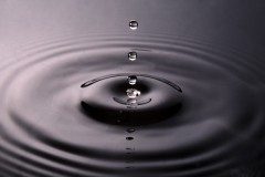 Macro shot of a clear water droplet above concentric ripples on a dark pool
